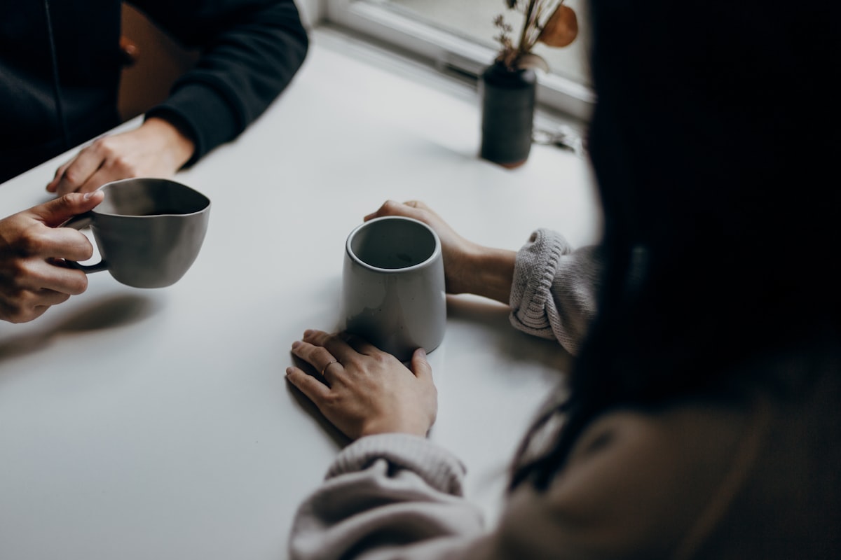 Dos personas conversando con café en una mesa junto a la ventana