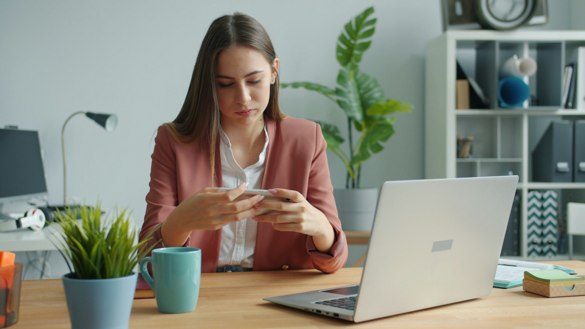 Mujer revisando su teléfono junto al notebook en el escritorio